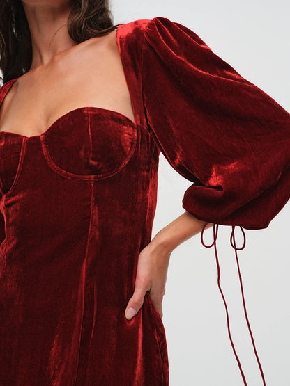 Woman wearng a deep red velvet mini dress. The dress features a structured bust, long billowy sleeves with adjustable ties, and an open back with a clasp.